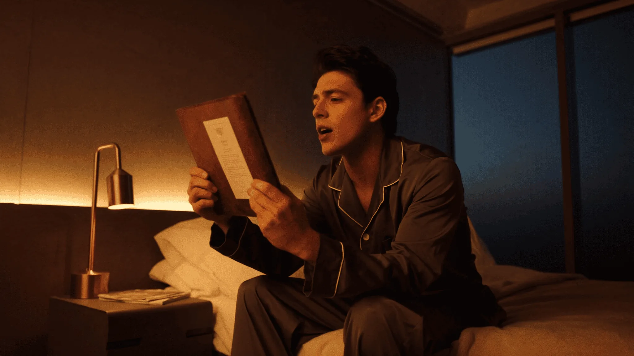 A young man on the edge of his bed in warm evening lamplight, reading his vision card aloud.